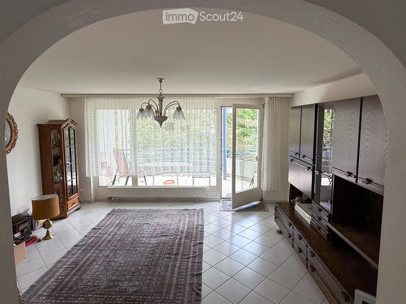 Bright living room with tiled floor, large window with sheer curtains, balcony access, chandelier, cabinet, table lamp, mirror, television, and area rug.