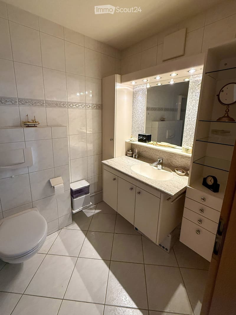 Modern bathroom with white tiles, a toilet, a sink with a mirror, and a cabinet. The sink has a modern faucet and a countertop with various toiletries. A small trash bin is placed next to the toilet, and a clock is visible on the cabinet.