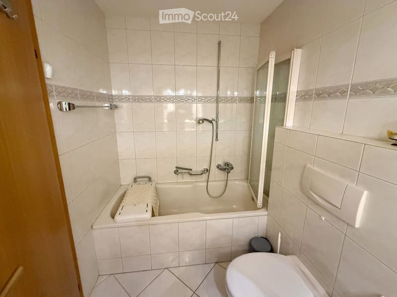 A small bathroom with a bathtub, toilet, and shower. The walls are tiled in light beige with a decorative border. A towel is placed on the edge of the bathtub, and a shower curtain is partially visible.