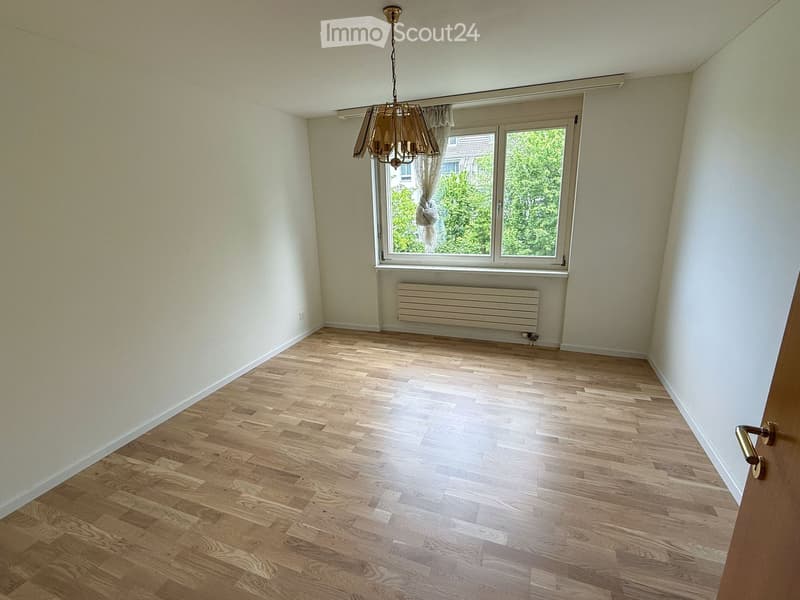 Empty room with wooden flooring, white walls, a window with curtains, a radiator, and a chandelier.