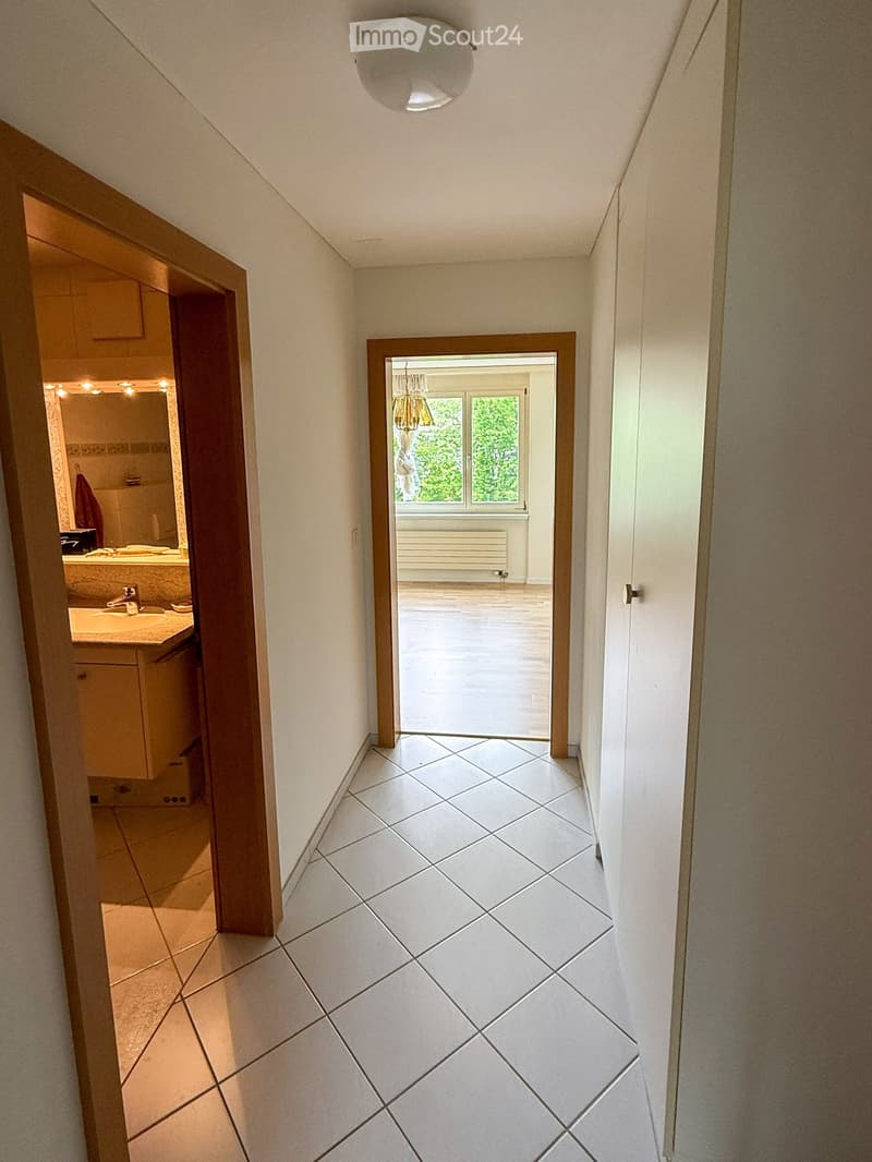 A narrow hallway with white tiled flooring, white walls, and a ceiling light fixture. The hallway leads to a bathroom on one side and an open area with large windows on the other.