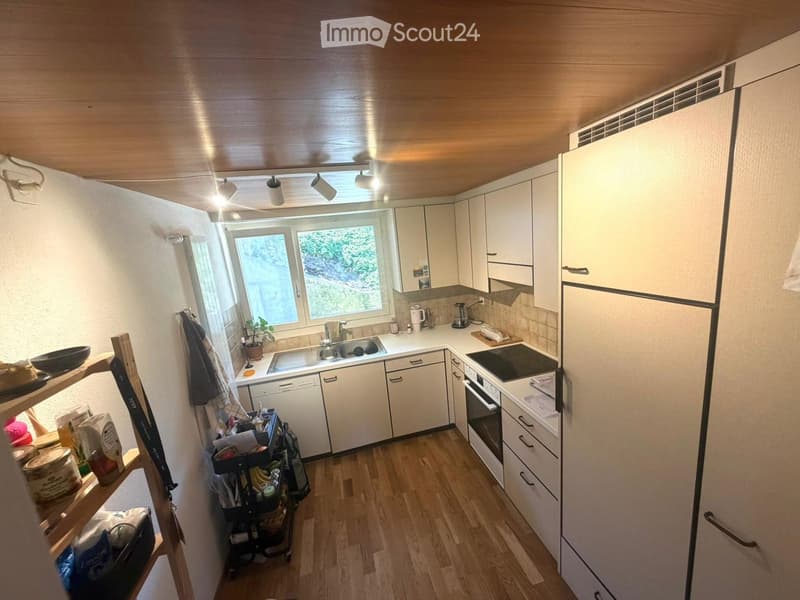Modern kitchen with white cabinets, wooden flooring, and stainless steel appliances including a refrigerator, oven, and dishwasher. A window provides natural light, and a shelf holds various items.