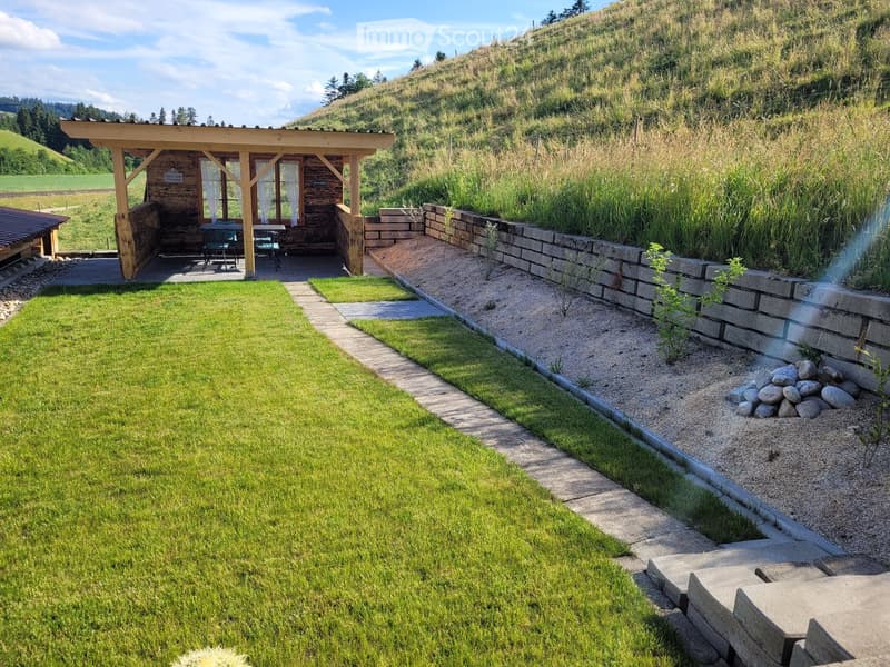 Well-maintained garden with a grassy lawn, stone pathways, a raised wooden deck with outdoor furniture, and a stone retaining wall with gravel and small plants.