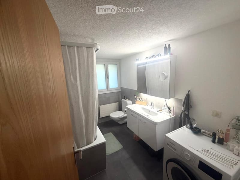 Modern bathroom with a white sink, mirror, toilet, and washing machine. Features a shower area with a curtain, a window, and various bathroom accessories on the counter.