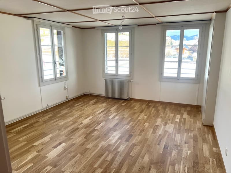 Empty room with wooden flooring, white walls, and large windows offering a view of the outside. A radiator is visible against one wall.
