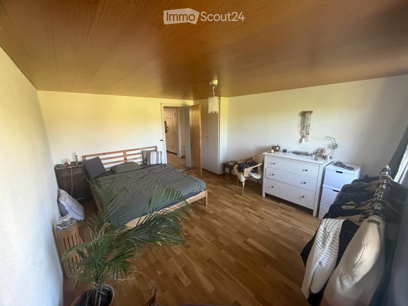 Cozy bedroom with wooden flooring, a bed with grey bedding, a nightstand, a dresser, a chair, a plant, and a wardrobe with hanging clothes.