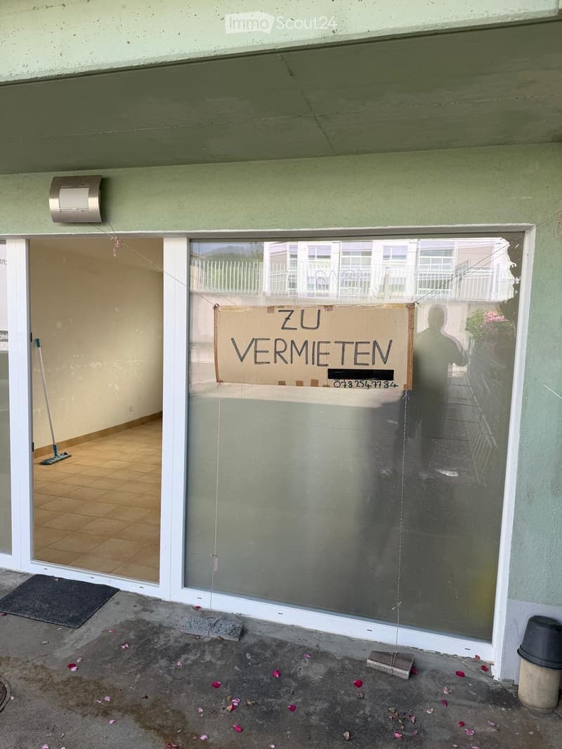 Empty room with tiled floor, white walls, and a broom leaning against the wall. A sign on the glass door reads "ZU VERMIETEN" (For Rent).