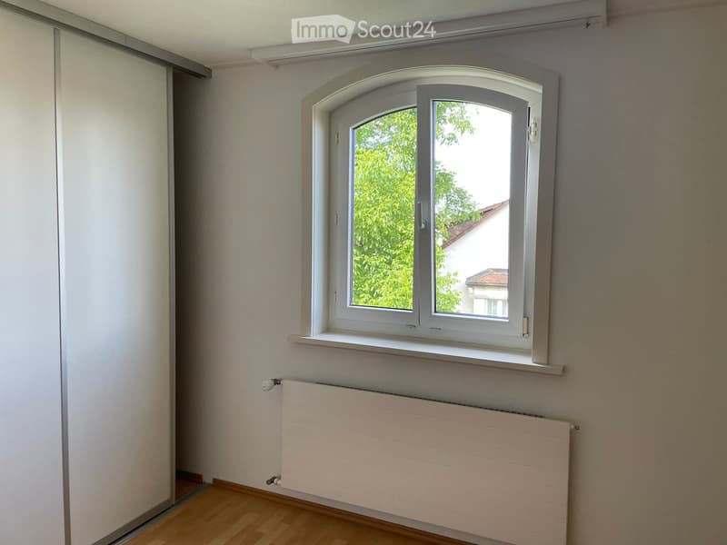 Empty room with light-colored walls, wooden flooring, a white radiator, and a large window with white frame and blinds.