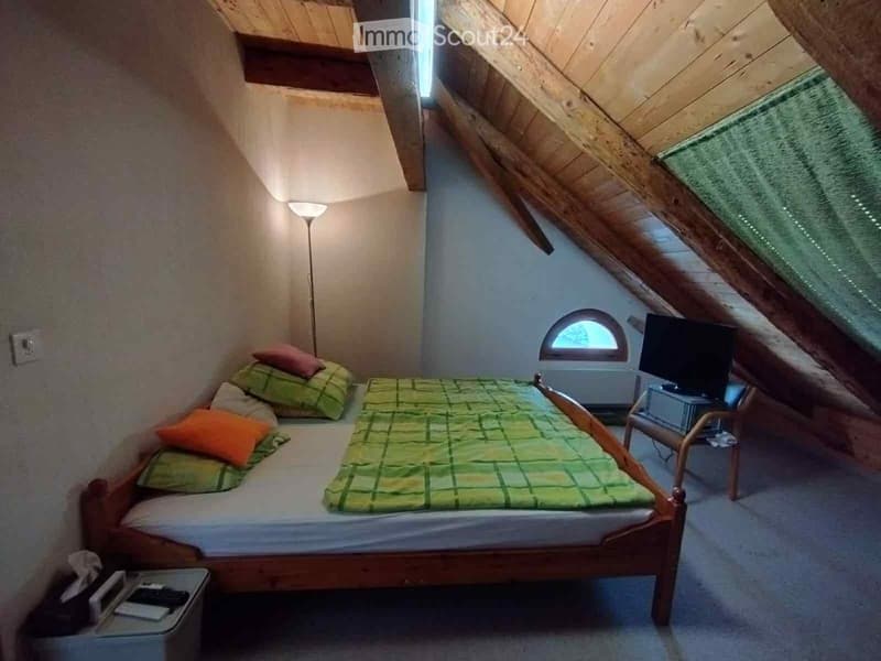 Cozy attic bedroom with wooden slanted ceilings, a double bed with green and orange bedding, a small chair, a table with a lamp, and a TV screen on a stand.