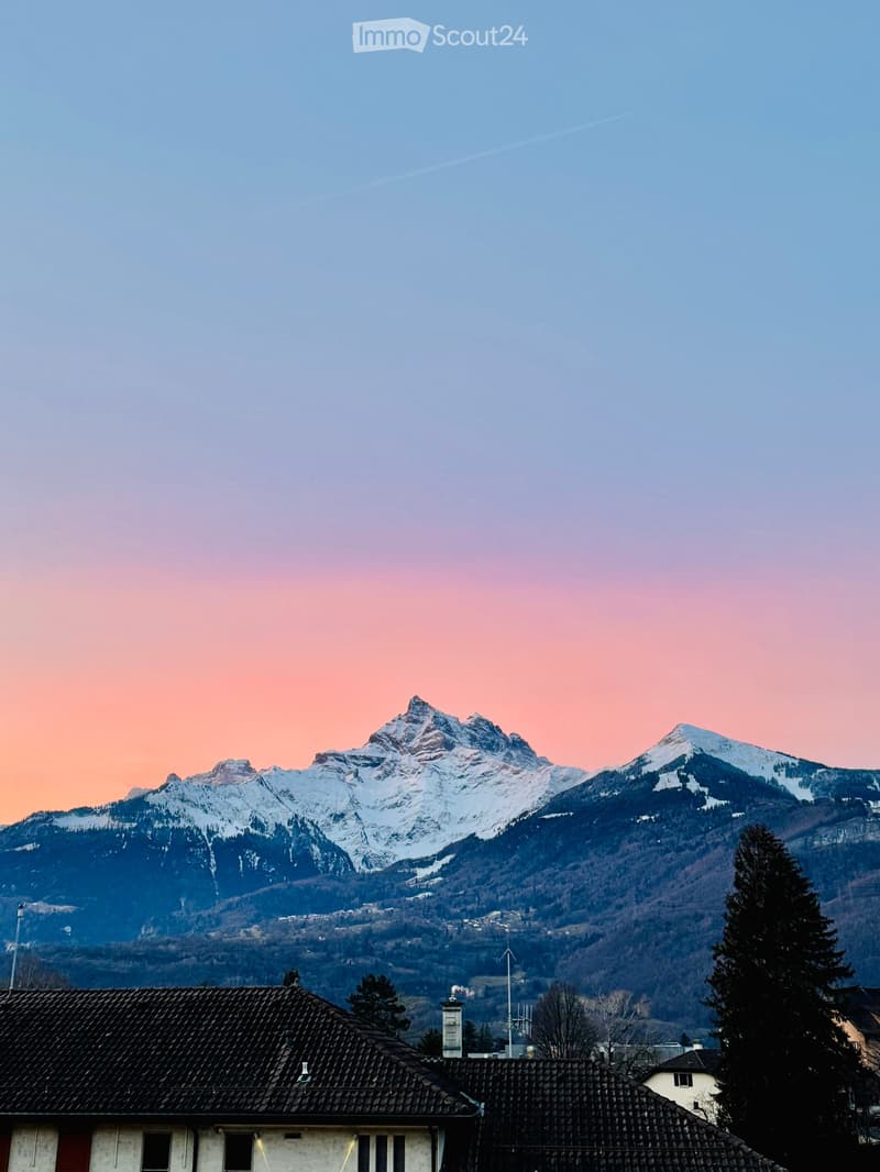 A house with a tiled roof and chimney, situated in a valley with snow-capped mountains in the background during sunset.
