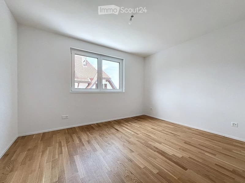Empty room with wooden flooring, white walls, and a large window showing a roofed building outside.