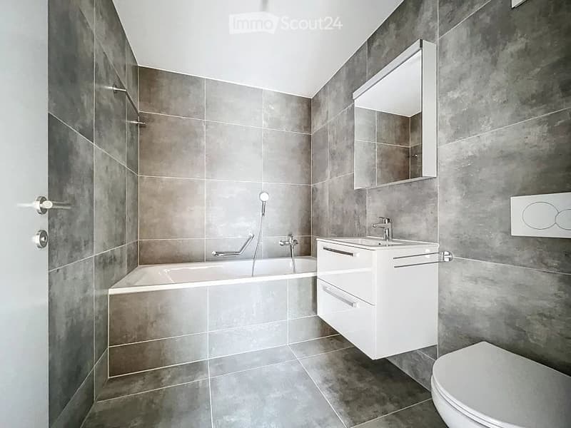 Modern bathroom with grey tiled walls and floor, featuring a bathtub, a white sink with a mirror cabinet, and a toilet.