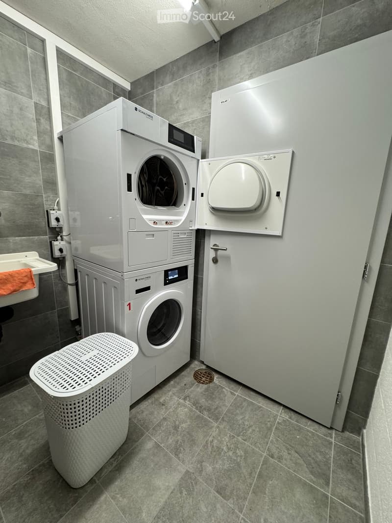 Modern bathroom with a washing machine, dryer, sink, and laundry basket. The room features grey tiled walls and floor.