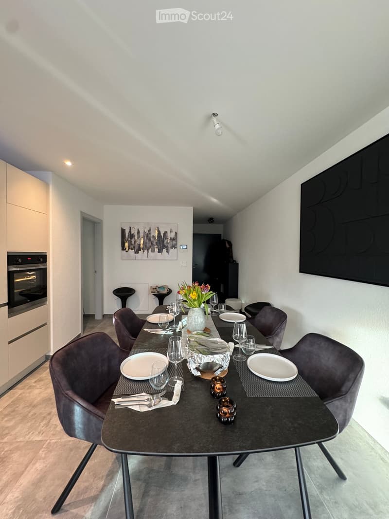 Modern dining area with a round black dining table, six dark grey chairs, a vase with flowers, and a small oven in the background.