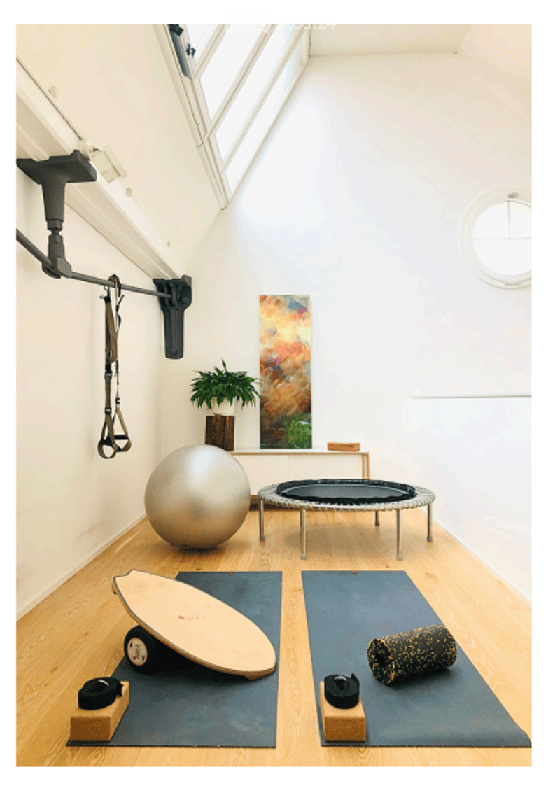 A modern, well-lit room with wooden flooring and white walls, featuring a fitness ball, yoga mats, a skateboard, a small table with a plant, and a colorful artwork on the wall.