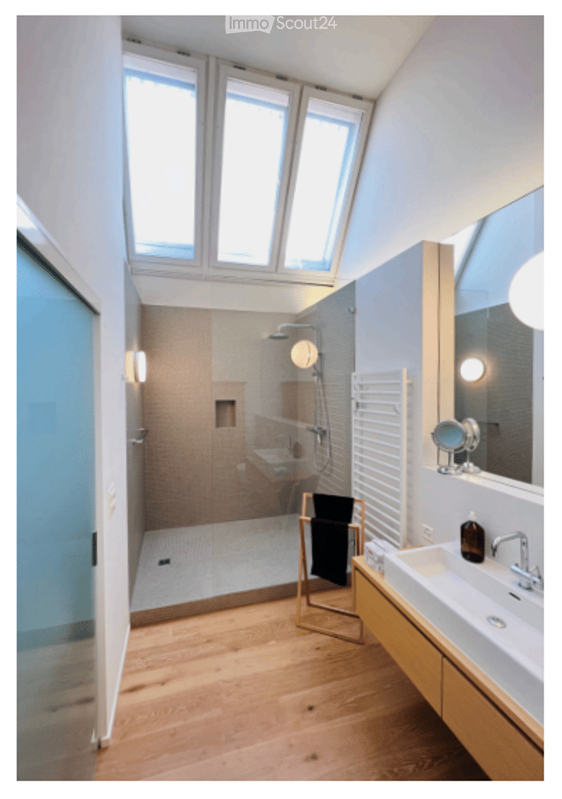Modern bathroom with wooden flooring, large windows, a glass-enclosed shower, a sink with a mirror, and a towel rack with a black towel.