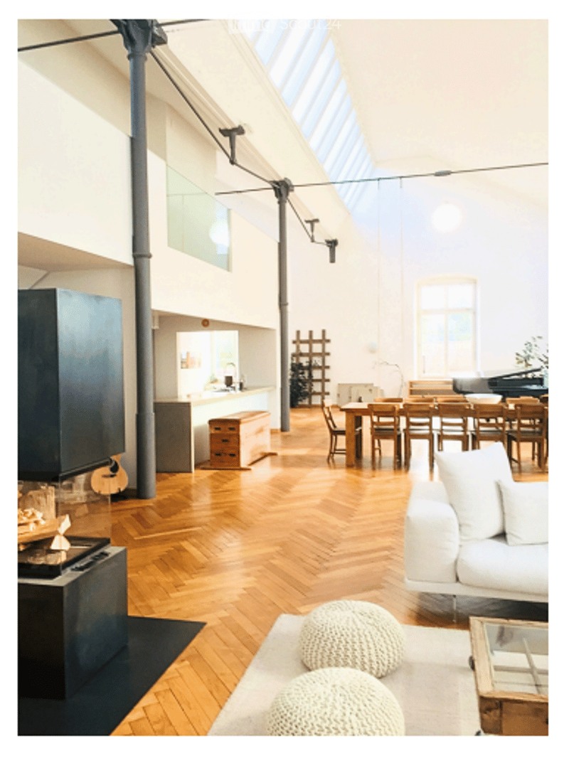 Open-plan living and dining area with wooden flooring, white walls, large windows, and skylights. Features a white sofa, coffee table, dining table with chairs, and decorative stools.