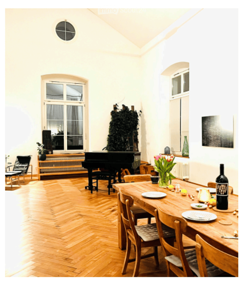 A spacious, well-lit room with wooden flooring, a grand piano, a dining table set for a meal, and several chairs around it. The room features large windows, a radiator, and a decorative plant in the corner.