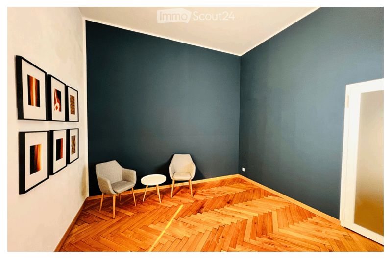 A small, modern room with dark blue walls and a herringbone wooden floor. Two light grey chairs and a small round table are placed near the wall. Several picture frames are mounted on the wall.