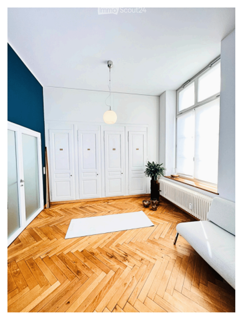 Empty room with wooden herringbone flooring, white built-in cabinets, large window with radiator, white sofa, small plant, and a white rug.