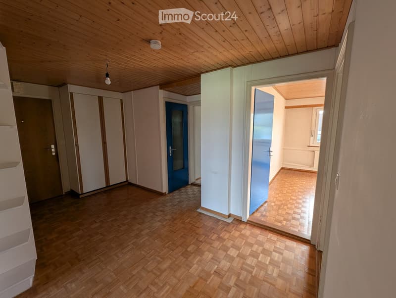 empty room, wooden parquet floor, wooden ceiling, white walls, two blue doors, white shelves, white cabinets, light bulb hanging from ceiling