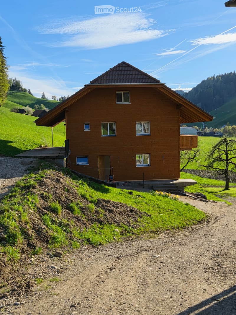 A wooden house with a sloped roof, multiple windows, and a small balcony, situated in a green, hilly landscape.