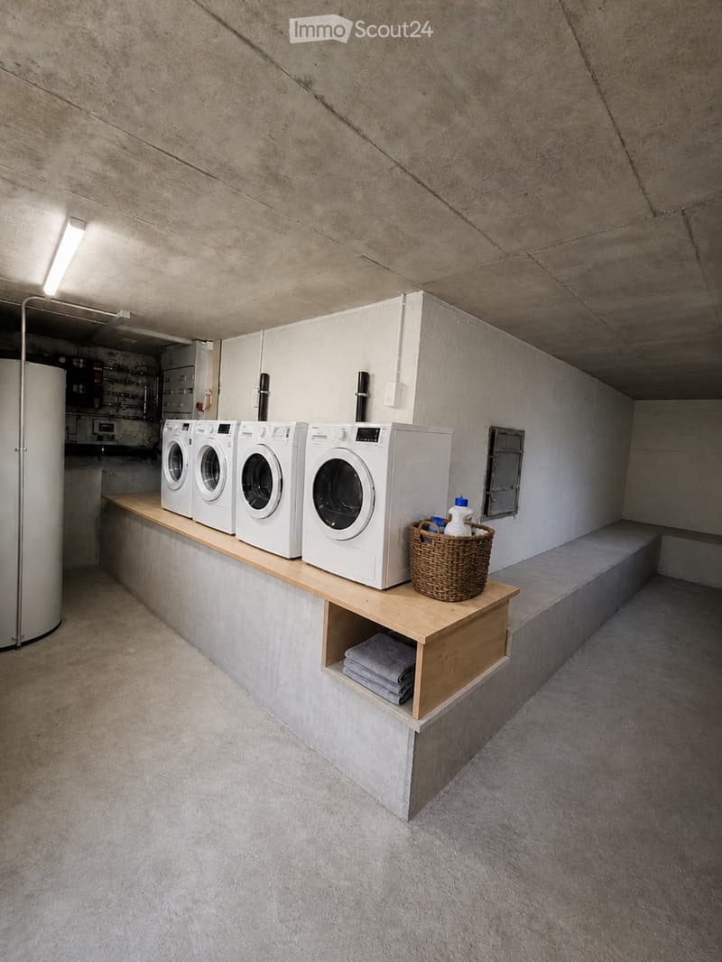 A modern, minimalist laundry room with four white washing machines and dryers arranged on a wooden shelf. A wicker basket with cleaning supplies sits on the shelf, and folded towels are stored in a compartment below.