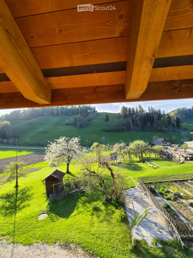 View from a wooden balcony overlooking a lush green garden with trees, a small shed, and a stone fence. The landscape includes rolling hills and a distant house.