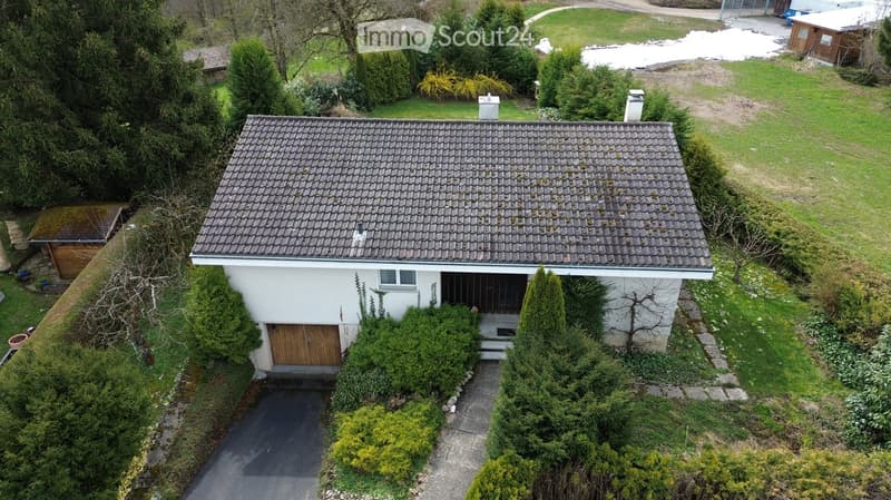 Aerial view of a single-story house with a tiled roof, white exterior, and a wooden garage door. The house is surrounded by a well-maintained garden with various trees and shrubs, and a paved driveway leads to the garage.