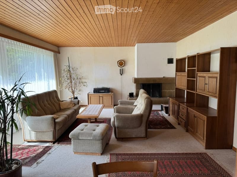 Spacious living room with wooden ceiling, fireplace, large windows, beige sofa, coffee table, ottoman, armchairs, bookshelf, and potted plants.