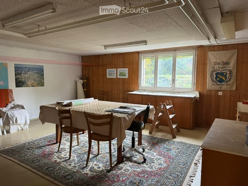 Room with wooden walls, a large window, a patterned rug, a table with chairs, a stool, a cabinet, and artwork on the walls.