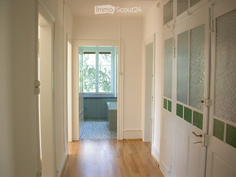 Empty hallway with wooden flooring, white walls, and multiple doors leading to other rooms. A small room with a window and tiled floor is visible at the end.