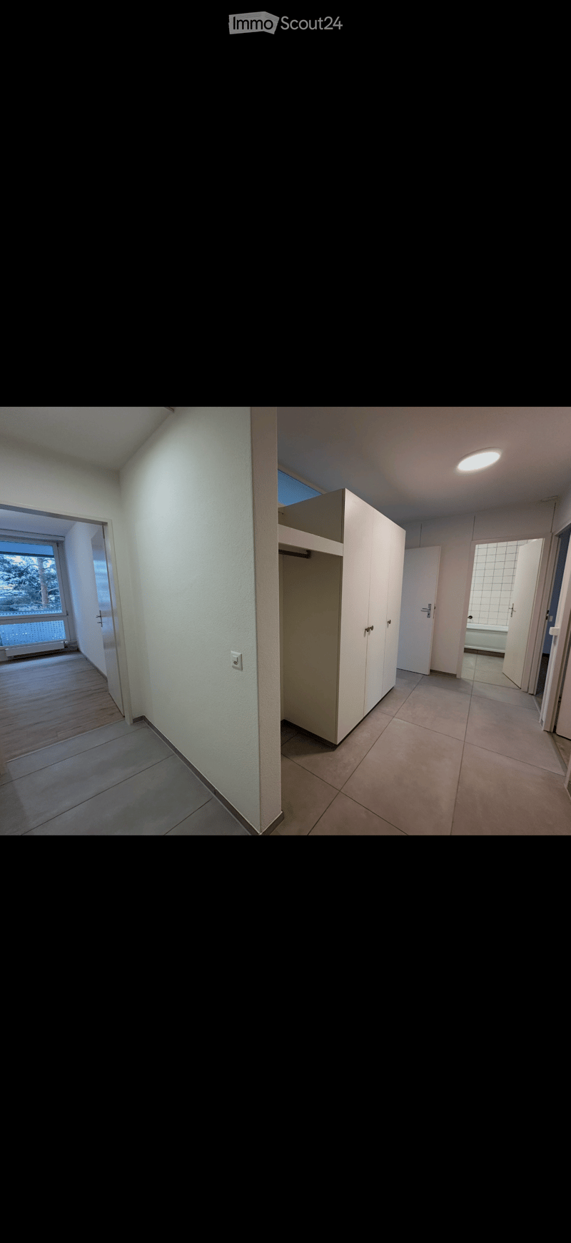 Empty hallway with tiled floor, white walls, and a built-in closet. The hallway leads to a room with a window and another room with a bathtub visible through an open door.