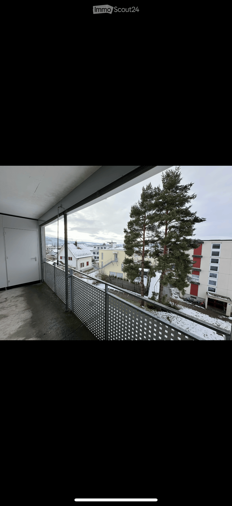 Balcony with metal railing, overlooking a snowy landscape with trees and neighboring buildings.