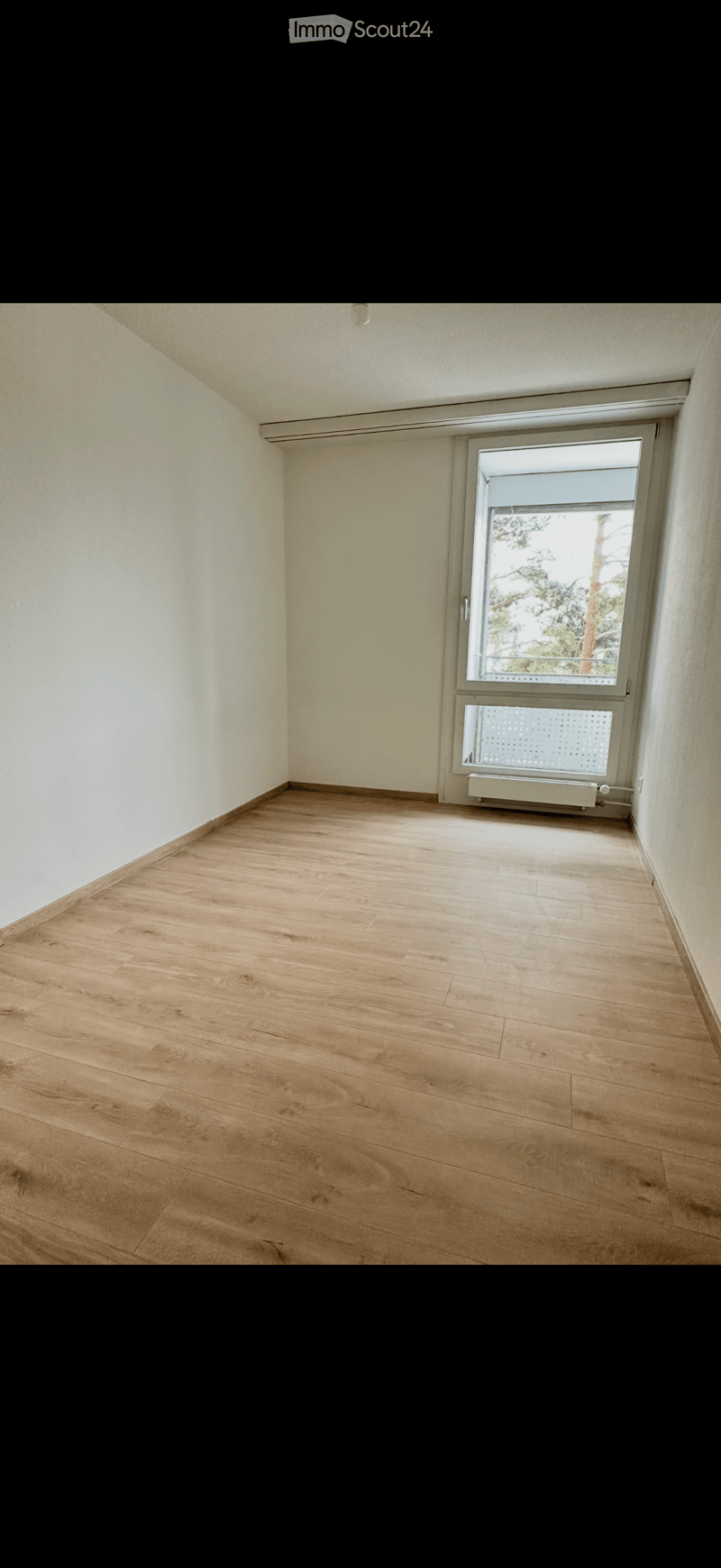 Empty room with light wooden flooring, white walls, and a large window with a view of trees.