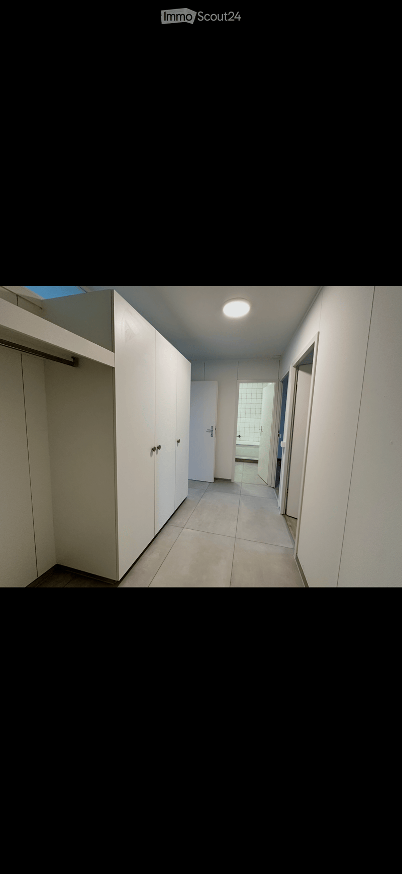 Empty hallway with white walls, tiled floor, and a ceiling light. Doors lead to adjacent rooms, including a bathroom visible at the end.