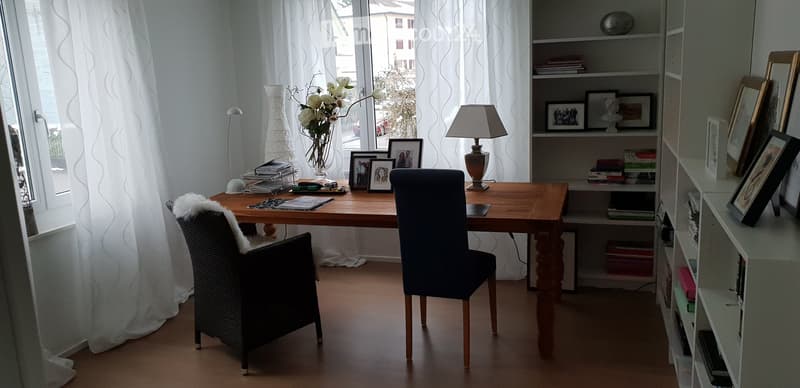 A well-lit study or home office with a wooden desk, two chairs, a bookshelf filled with books and decorative items, a table lamp, and a vase with flowers on the desk.