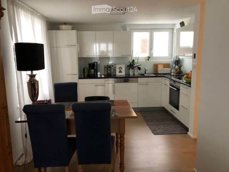 A modern kitchen and dining area with white cabinets, a wooden dining table with four chairs, a lamp, and various kitchen appliances.