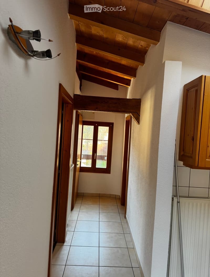 A narrow hallway with tiled flooring, wooden doors, and exposed wooden beams on the ceiling. A window at the end provides natural light, and a radiator is visible on the right side.