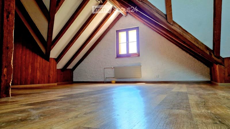Empty attic room with wooden beams, wooden floor, white walls, and a small window with purple frame.