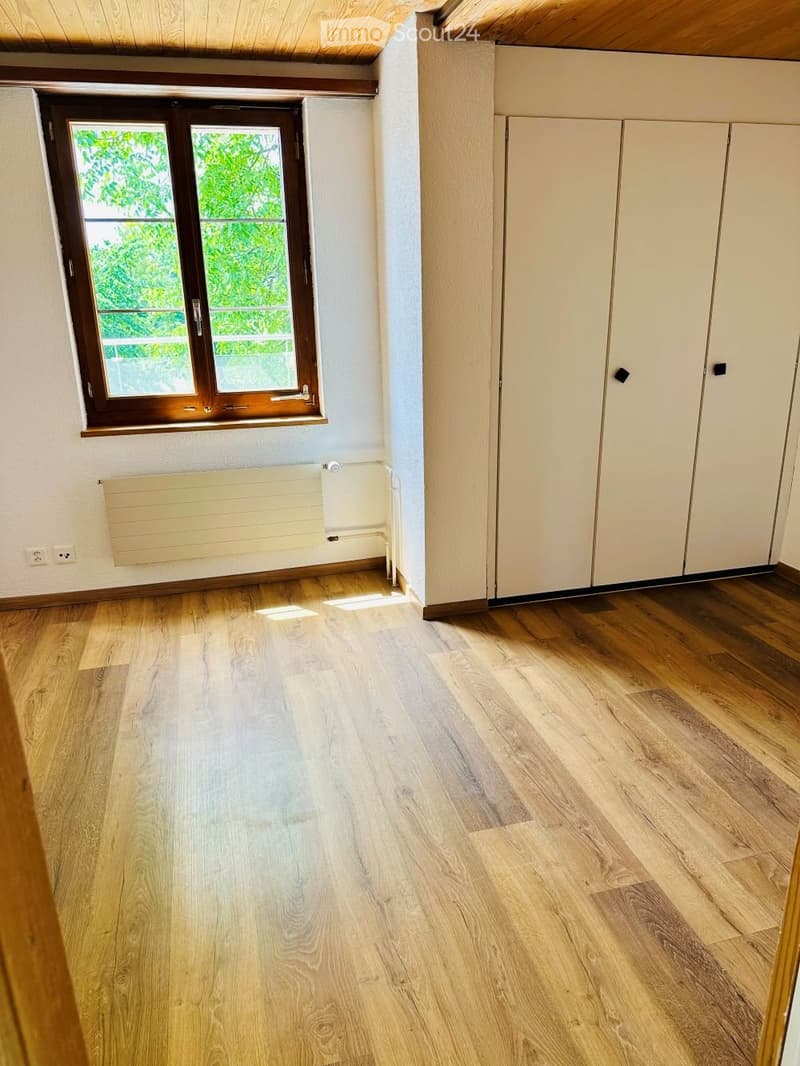 Empty room with wooden flooring, white walls, built-in closet, window with wooden frame, radiator, and electrical outlets.