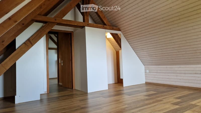 Empty attic space with wooden beams, wooden flooring, white walls, and a wooden door leading to another area.