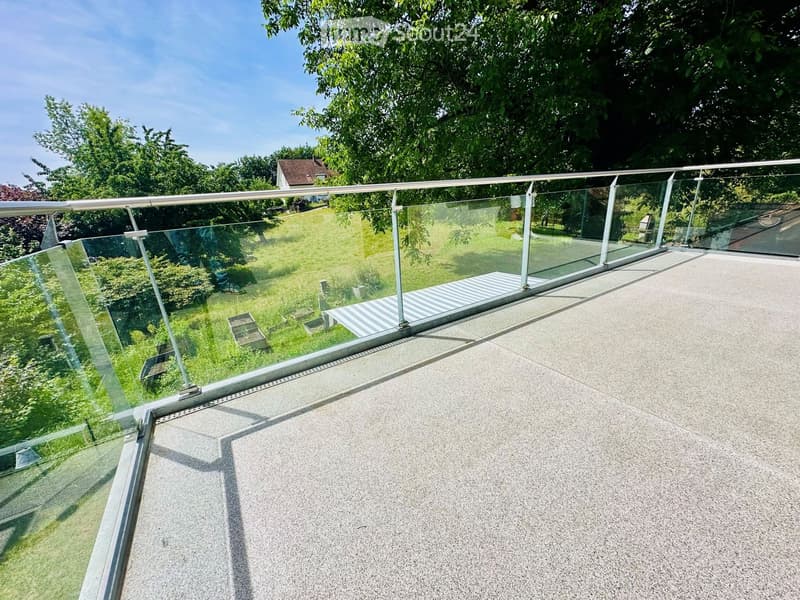 Modern balcony with glass railing, concrete floor, and view of a green lawn and trees.