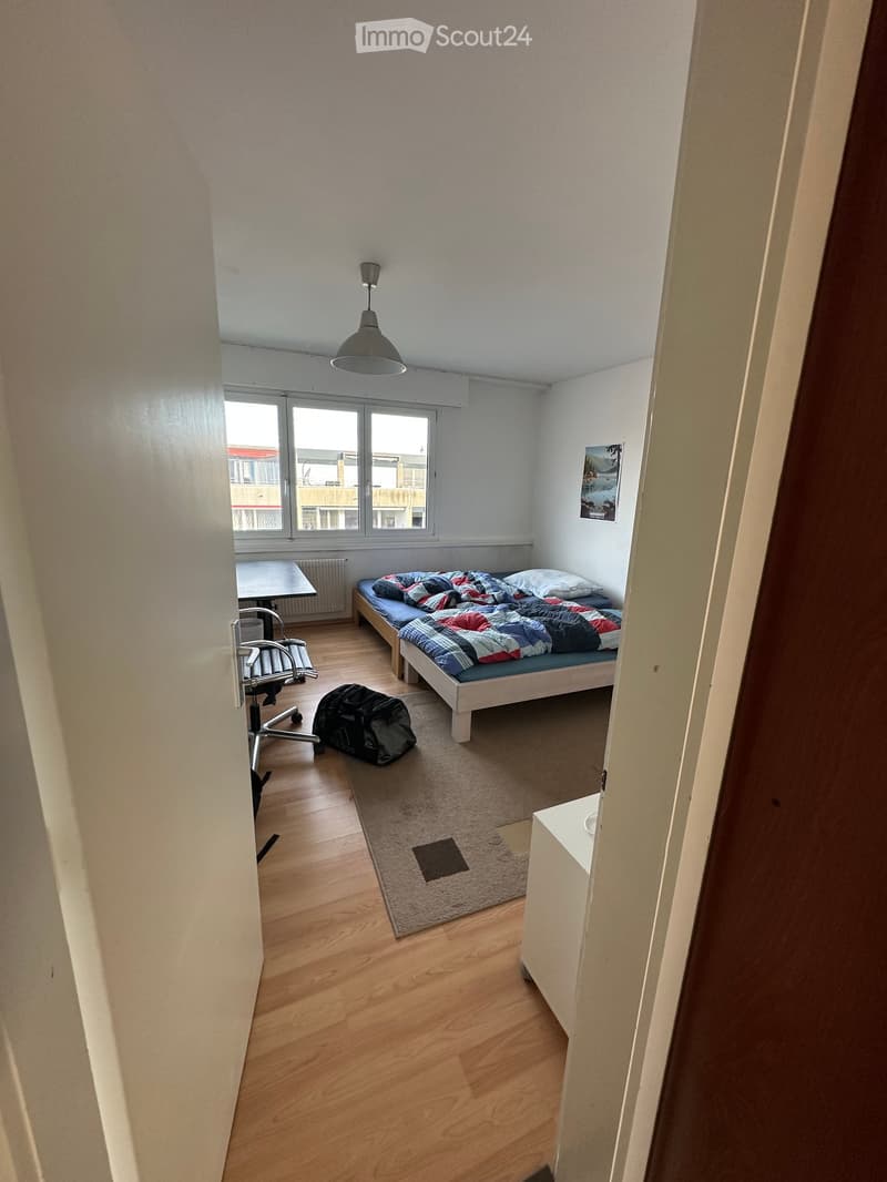 A small, furnished bedroom with a single bed, a chair, a table, a backpack, and a picture frame on the wall.