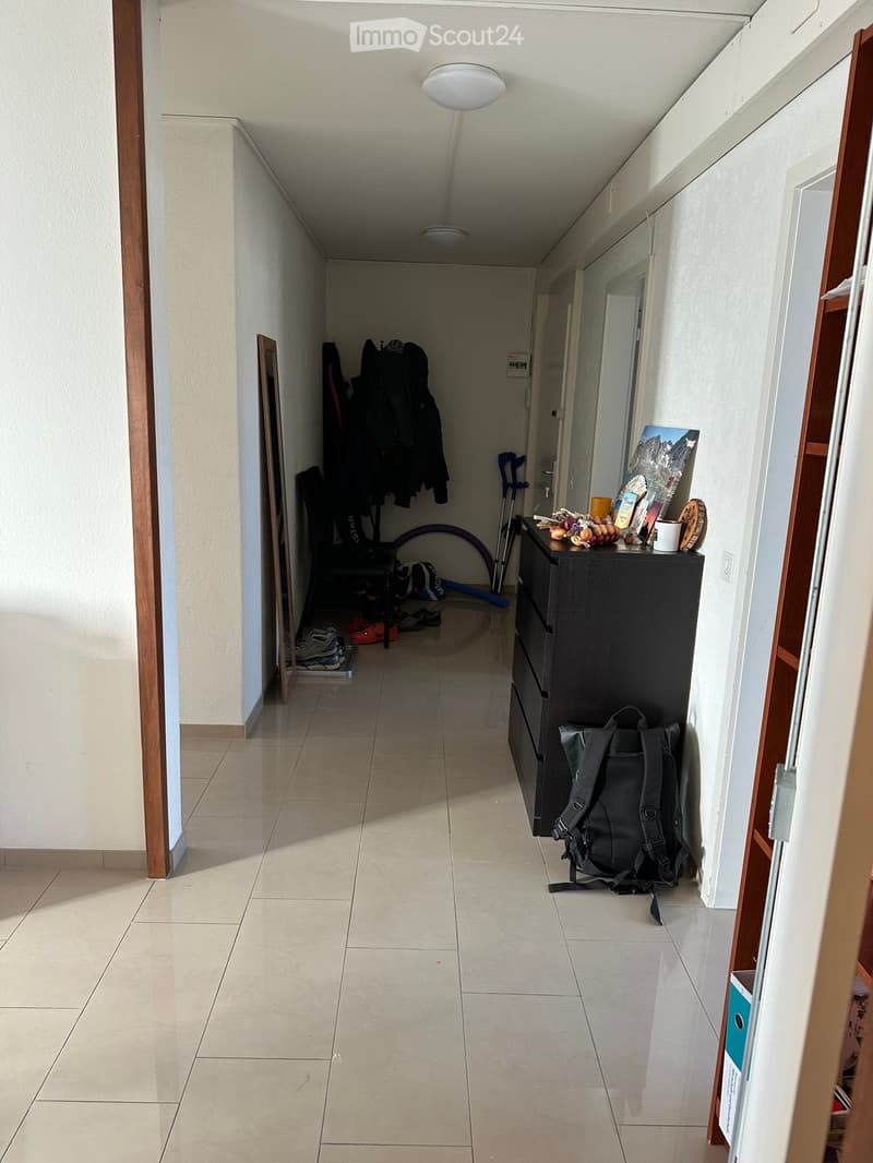 A narrow hallway with tiled flooring, white walls, and wooden trim. Visible items include a black dresser with a backpack, shoes, and a coat rack with jackets and shoes.
