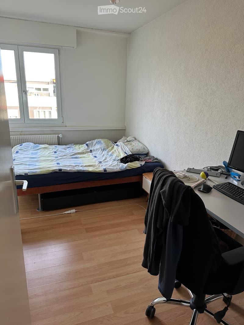A small bedroom with a single bed, a desk with a computer, and a chair. The room has wooden flooring and large windows letting in natural light.
