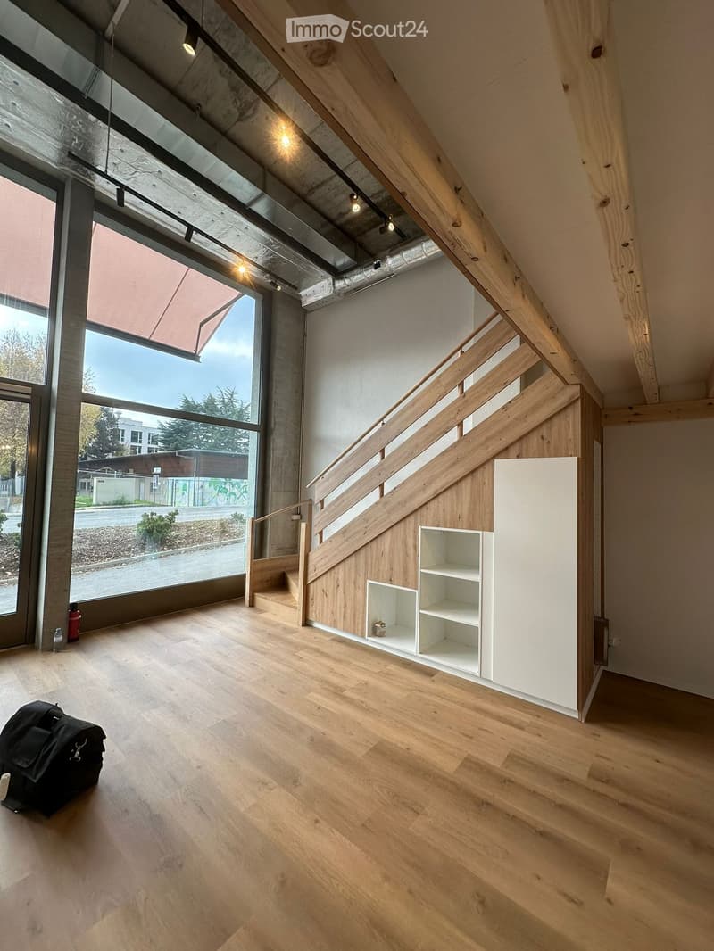 Empty room with wooden flooring, white walls, a staircase with wooden railings, and large windows offering a view of the outside. A small built-in shelf unit is visible on one wall.