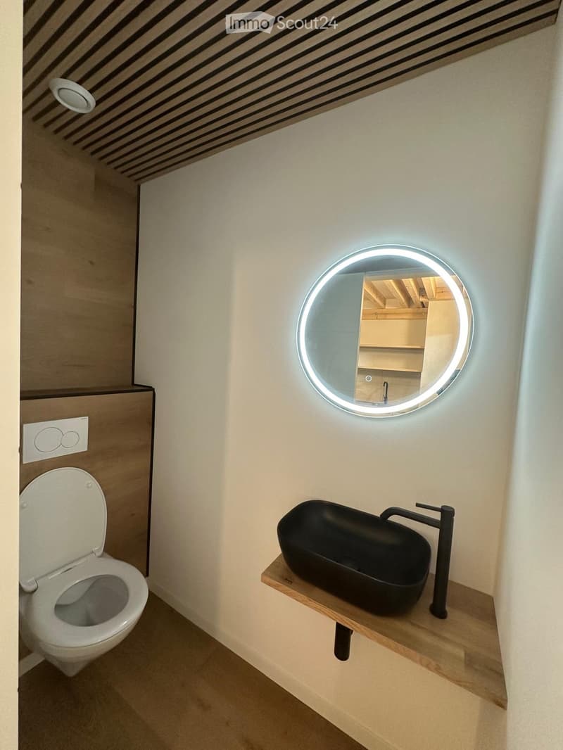 Modern bathroom with a sleek black sink, wooden shelf, round illuminated mirror, and a toilet.