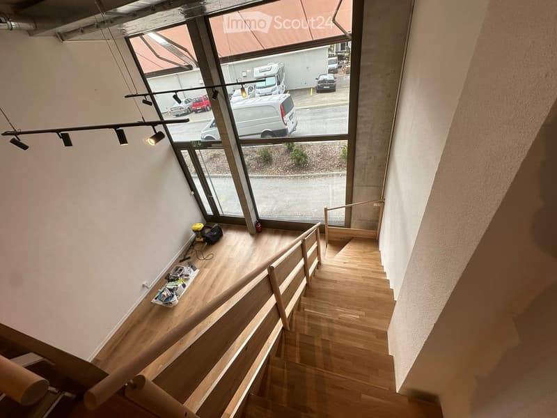 Staircase with wooden steps and railings, large windows overlooking a parking area with several cars, and some tools and equipment placed on the landing.