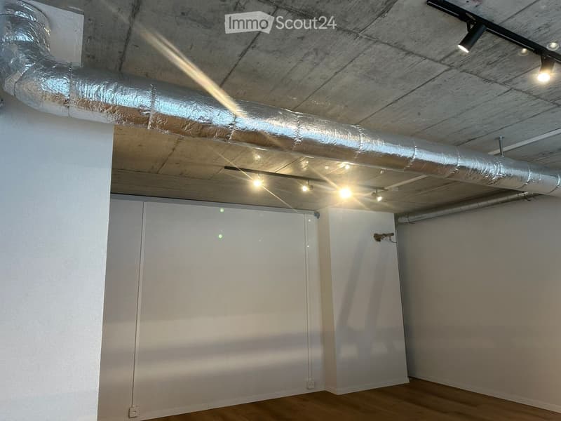 Empty room with wooden flooring, white walls, and exposed wooden ceiling with lighting fixtures. There are two doors and visible air ducts running across the ceiling.